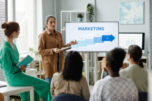 Young businesswoman standing near monitor with graphs at seminar at office and presenting new marketing strategy to colleagues