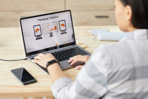 High angle view at young woman looking at laptop screen with Marketing Statistics headline while sitting by wooden table and working at home office, copy space